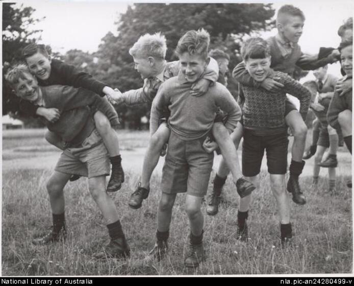 A group of young boys in a field of grass. Three boys carrying another three boys on their backs, and are smiling and reaching out to each other.