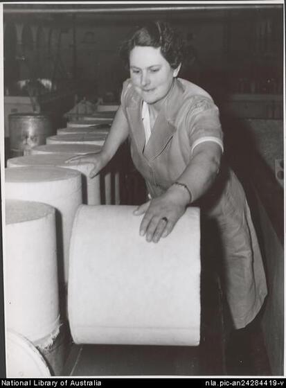 A middle aged woman wearing a short sleet coat leans over and rolls a large cylinder of cheese. To her right is a row of large cheese cylinders on their ends.