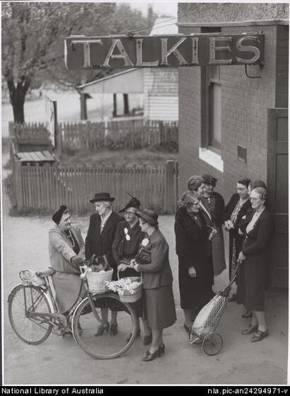 A group of women wearing coats, skirts and hats stand in two smaller groups in front of a brick building to the right with a sign above reading 'Talkies'. One woman has a bicycle, two carry baskets, and one holds a two-wheeled bag. 