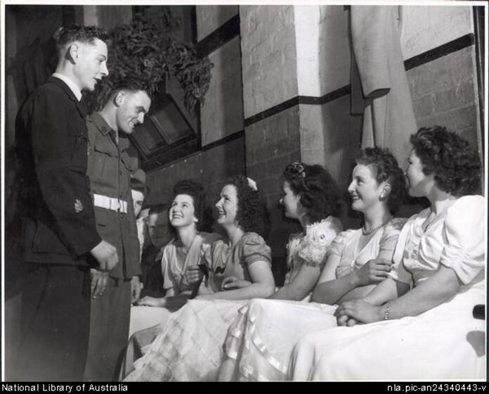Two young men in military uniform stand looking down at a row of five seated young women wearing light dresses. All are smiling.