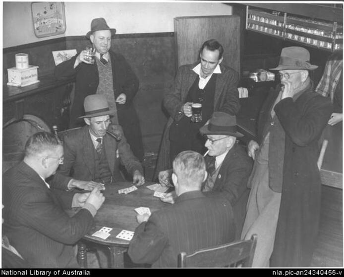 A group of four men wearing suits and hats seated around a circular table with playing cards on the surface. Two men stand behind drinking, and a third stand holding a cigarette to his mouth. 