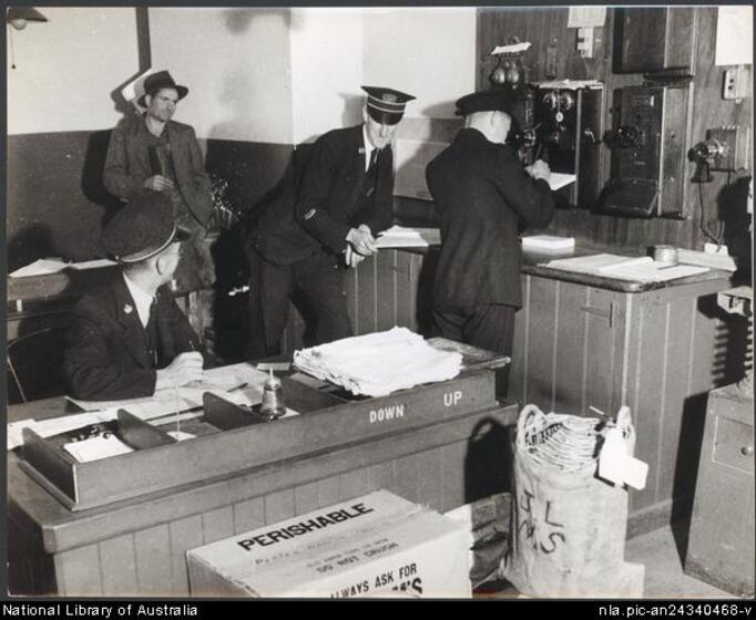 A man in uniform and hat is seated at a desk with a mail tray, looking back at another two men in uniform, one leaning on a bench. Behind is a fourth man in a jacket and hat. In the foreground are boxes and a mail box. On the wall at the back are old style telephones and receivers.