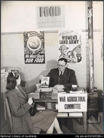 A young woman sits in front of a desk and hands a book titled 'Food Front' to an older man behind the desk. A sign on the front of the desk reads 'War agricultural committee'. Three posters are behind reading 'We must share our food', 'Food, One weapon Australia is pledged to supply' and 'A vital war job, join the women's land army'. 