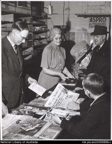 Four men in suites and hats and one woman in a dress stand around and look down at a central long table with newspapers spread out on top.  Shelving is behind.
