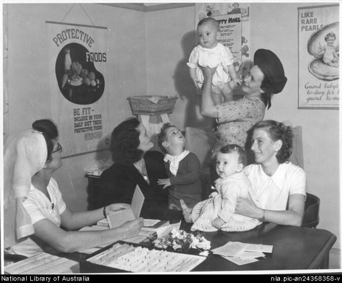 A woman wearing a nurses' hat and white short sleeved top is seated at a table holding a booklet, looking up at a infant child held up by a young woman in a floral dress and hat. Two other young women sit behind the table, each holding an infant child. 