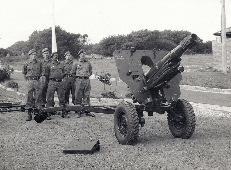 Five men in military uniform and beret bats stand behind a large metal gun on wheels. A flagpole and shrubs are seen behind.