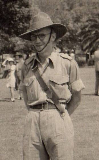 A man wearing short sleeved military uniform, glasses, and an Australian slouch hat, standing with arms behind his back. A palm tree, grass and other people can be seen behind.
