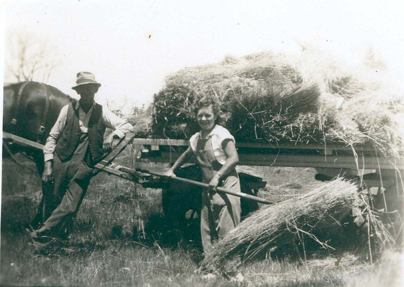 Young woman in overalls holding hay on a pitchfork in front of a cart of hay pulled by a horse. A man in a vest, trousers, shirt and hat stands leaning against the cart.