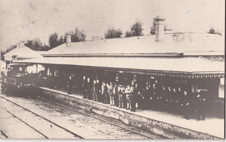 Sepia coloured photograph of country railway station with a train arriving, with two rows of people standing on the platform. 