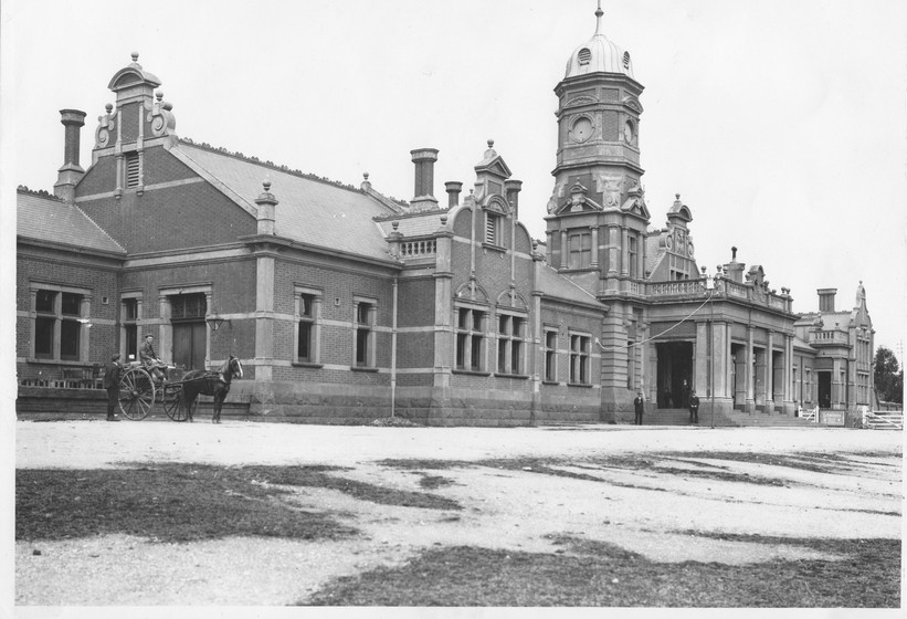 Landscape view of a large ornate train station front with clock tower in the centre. A horse and cart with two men can be seen to the right. 