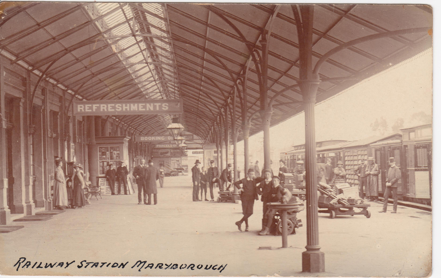 Train station platform with people in 1900s attire throughout. Platform has a pitched tin roof held up by columns, and a train at the platform to the right. A 'refreshments' sign is visible, and handwritten on the image is 'railway station Maryborough'.