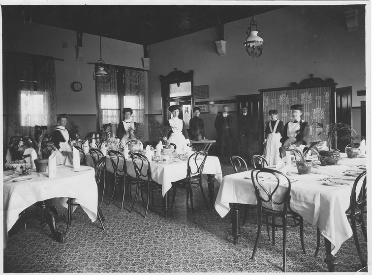 Three tables with white table cloths and set with dinner settings and potted plans, in a room with a patterned floor and with men and women in service uniforms behind.