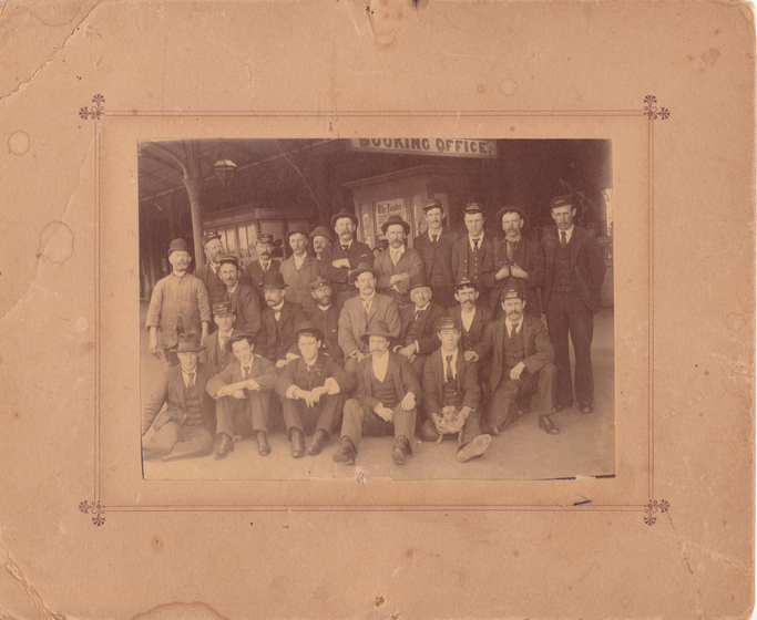 Group photograph mounted on card to form a large border. Images shows three rows of men in suites wearing hats in front of a building with a sign saying 'booking office' overhead. . Front row is seated.