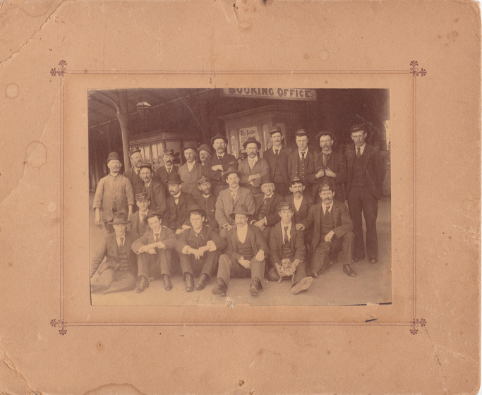 Group photograph mounted on card to form a large border. Images shows three rows of men in suites wearing hats in front of a building with a sign saying 'booking office' overhead. . Front row is seated.