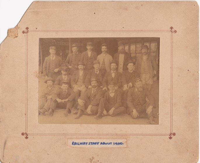 Group photograph mounted on card to form a large border. Images shows three rows of men in suites wearing hats in front of a building with a sign saying 'booking office' overhead. Front row is seated. Handwritten beneath is text 'railway staff about 1900'.