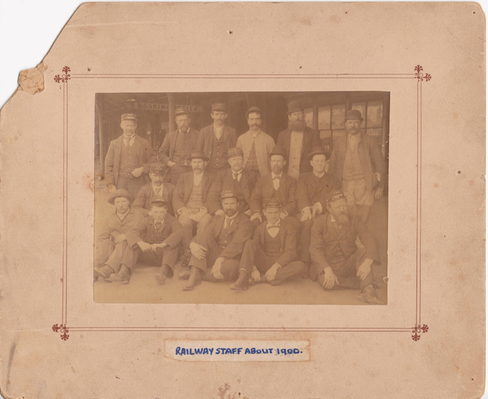 Group photograph mounted on card to form a large border. Images shows three rows of men in suites wearing hats in front of a building with a sign saying 'booking office' overhead. Front row is seated. Handwritten beneath is text 'railway staff about 1900'.