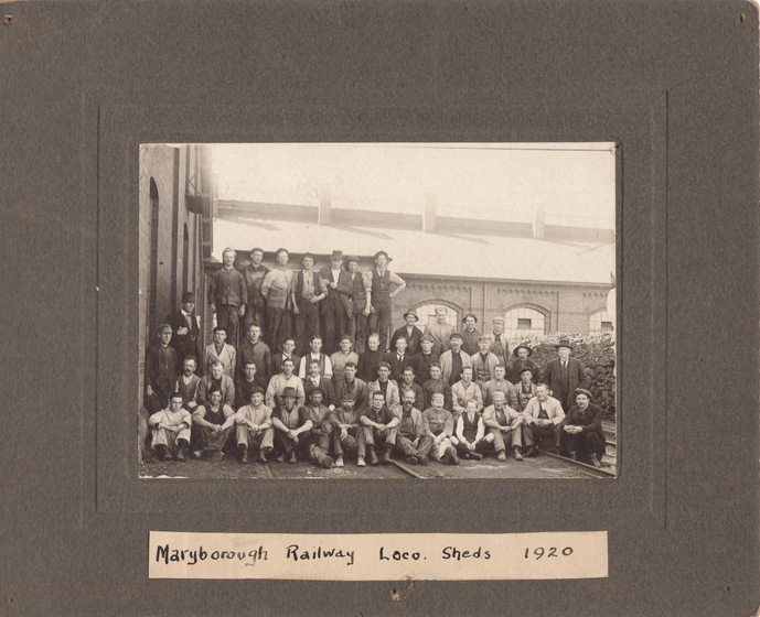 Group photograph mounted on dark card to form a large border. Image shows four rows of men in suites wearing hats in front of a building. The front row is seated on the ground, the back row is standing on a platform that is not visible. Handwritten beneath is text 'Maryborough Railway Loco Sheds 1920'