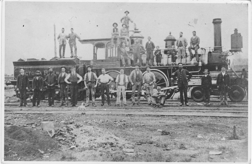 Train engine on a set of tracks, with men in work wear standing in front of, and on top of the engine.