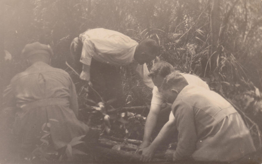 Blurry image of four men working in bushland wearing coats, crouched and leaning down over materials.