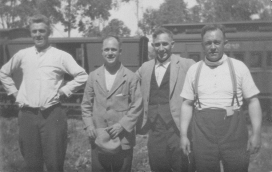 Four men standing together in work attire, in front of the carriages of a train with gum trees seen behind.