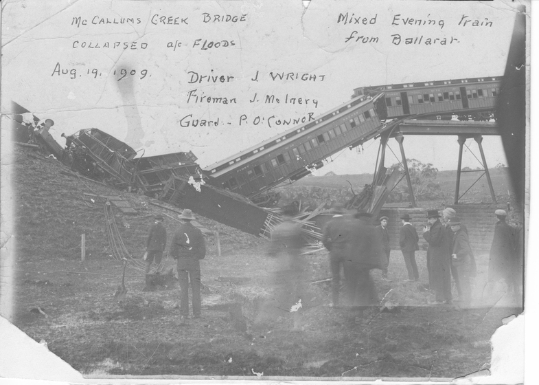 Photograph with bottom two corners torn off, of train carriages that have crashed, with a rail bridge partially fallen. People stand in the foregrown. Handwritten text on photograph reads 'McCallums Creek Bridge, Mixed Evening Train from Ballarat, Aug 19 1909. Driver J. Wright, Fireman J. McInery, Guard P.O. Connor'. 
