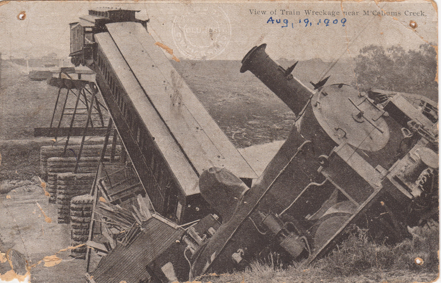 Train carriages that have crashed off a collapsed bridge, the train engine is seen falling back off a cliff in the foreround. Text above reads 'View of Train Wreckage near McCallum's Creek'. Handwritten text beneath reads 'Aug, 19, 1909'. 