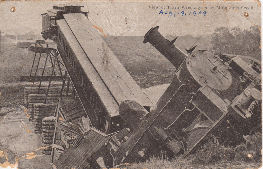 Train carriages that have crashed off a collapsed bridge, the train engine is seen falling back off a cliff in the foreround. Text above reads 'View of Train Wreckage near McCallum's Creek'. Handwritten text beneath reads 'Aug, 19, 1909'. 