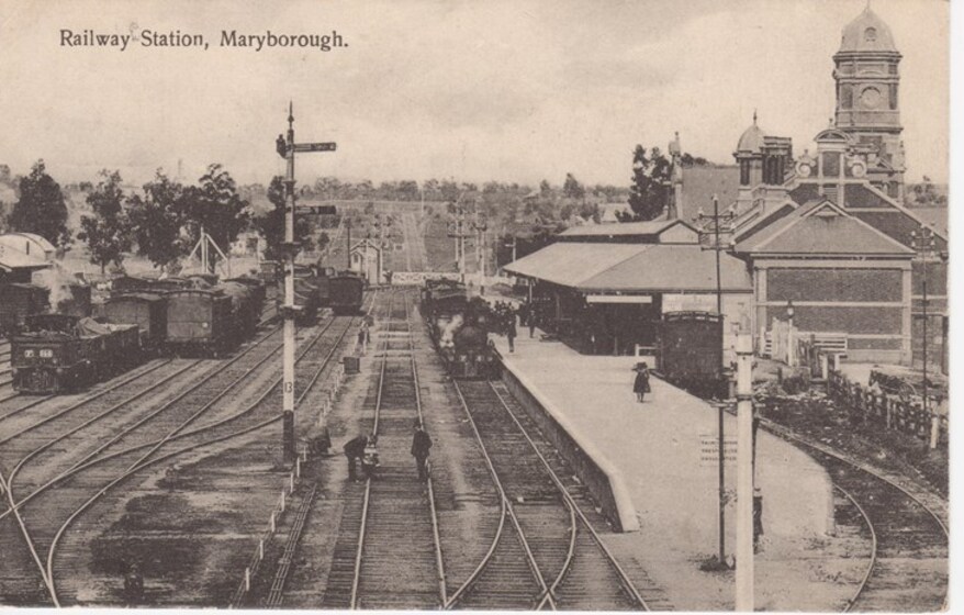 Landscape view of multiple parallel  train tracks with two people standing on the tracks. A train station is to the right with a train coming into the station, and carriages are seen on the tracks to the left. 
