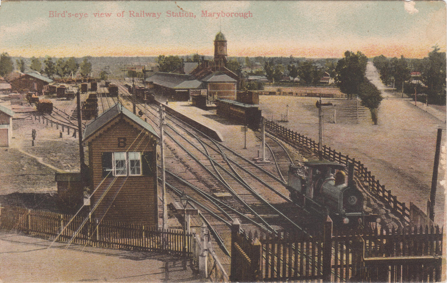 Arial view of a train engine in a small train yard, with a small brick building to the right, multiple parallel tracks, and the train station in the distance. Text on the image reads 'bird's eye view of railway station, Maryborough'. 