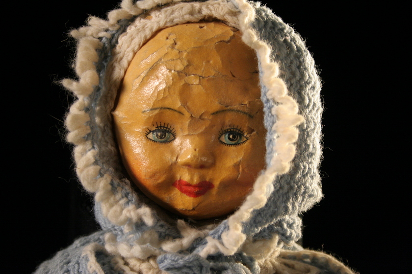 Head of a papier-mache doll with a light blue and white wool crochet hood on a black background.