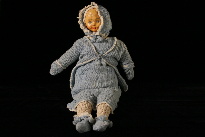 Doll with a papier-mache head wearing a light blue wool crochet dress, and hooded jacket, with cloth woollen legs showing on a black background. 