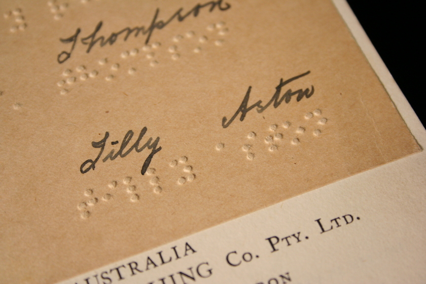 Close up view of a book page featuring handwritten text with braille underneath, text reads 'J Thompson' and 'Lilly Aston'.