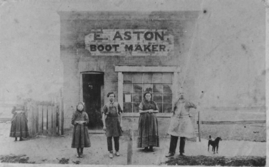 A girl, young man, woman and older man standing in front of a brick building with a black dog. The building reads 'E. Aston Boot Maker'. Another woman stands to the left edge beside the building.