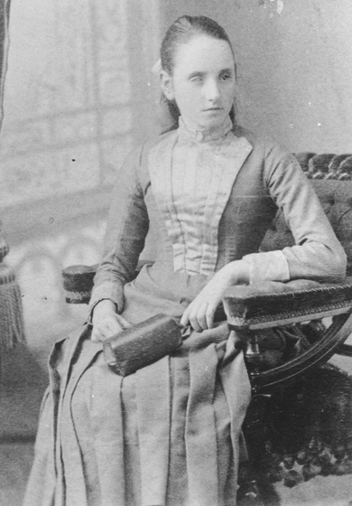 Young girl seated in an ornate chair, wearing an 1890s dress and holding a small bag.