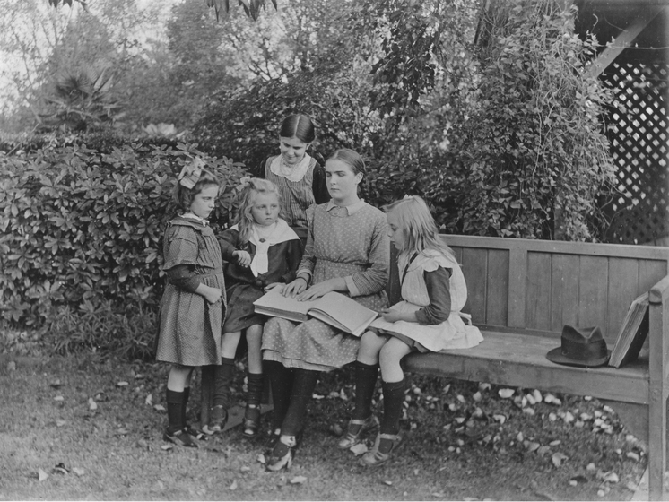 A young woman sits on a bench in a garden with a book in her lap and fingers on the page. She is surrounded by four young girls.