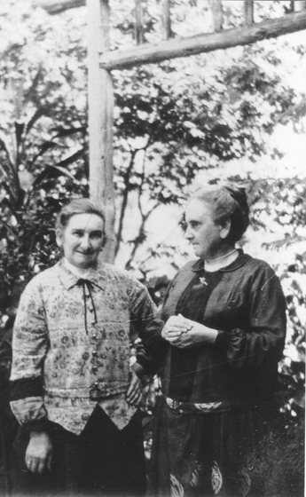 Two elderly women standing before trees. 