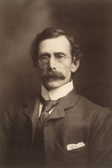 Torso studio portrait of a middle aged man with a moustache wearing glasses, a suit and tie.