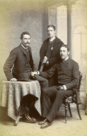 Three men in suites sit and stand around a small table with a lace table cloth. Two men have moustaches.