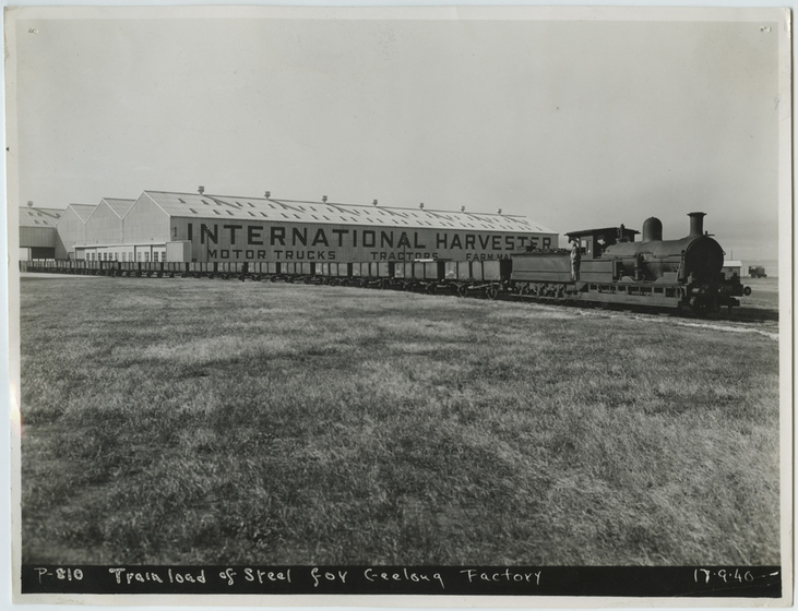 Train with carts seen travelling through a grass field. An industrial building behind features the words 'International Harvester, Motor Trucks, Tractors''