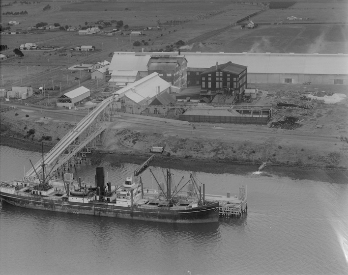 Ship in a body of water, docked at a wharf. Industrial area and buildings is seen behind on the bank.