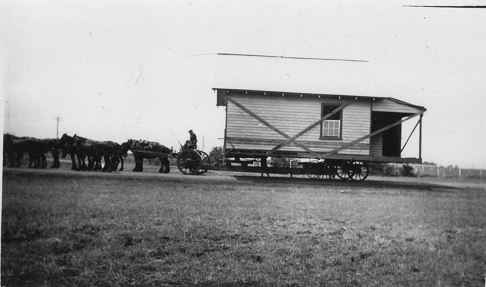 Part of a house mounted on cart wheels being pulled by horses through a field. 