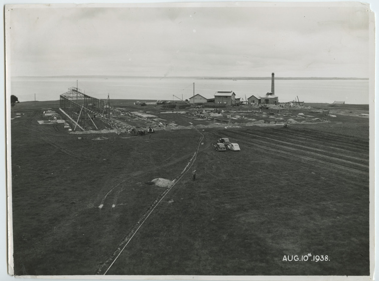 Aerial view of a field with an industrial site at the rear on the water front. Three cars can be seen in the mid-ground.