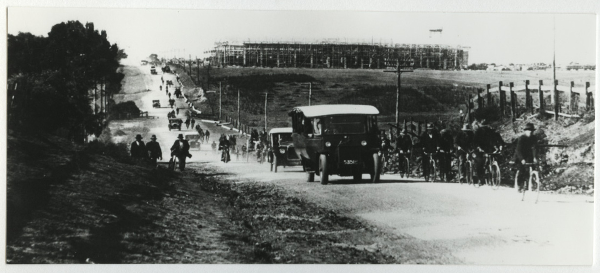 Dirt road with 1920s vehicles and men riding bicycles, both travelling away from a building under construction in the distance. 