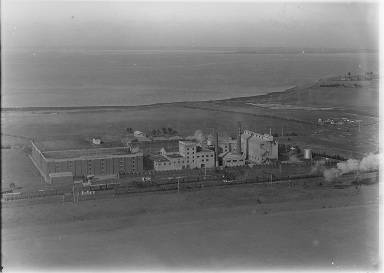 Aerial view of industrial buildings surrounded by grass fields, with the water front behind.