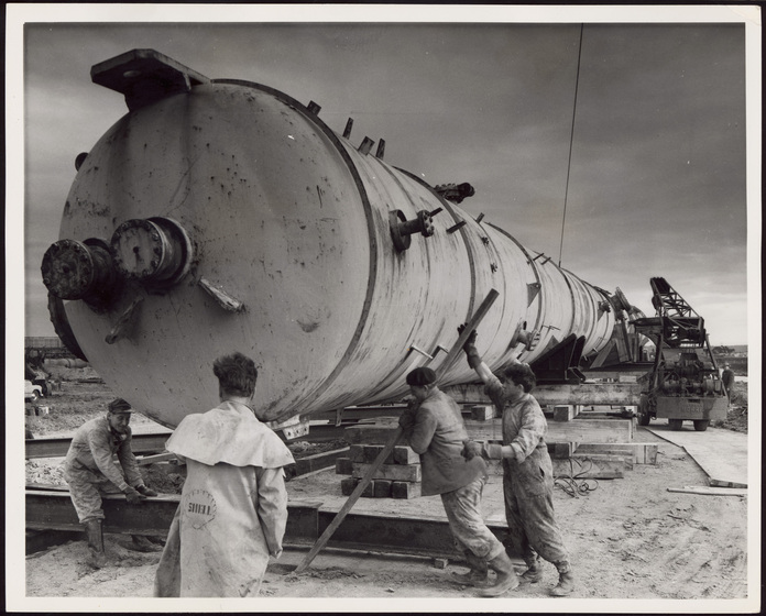 Four men in work wear stand around a large piece of cylindrical machinery. Two men are levering a piece of timber beneath the front. 
