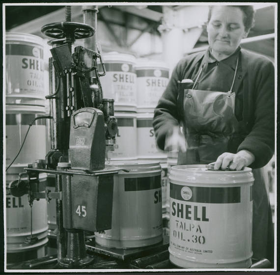 Middle aged woman wearing a plastic apron, standing behind and sealing a small metal barrel with the word 'SHELL' on the side. She is surrounded behind by other similar barrels.  