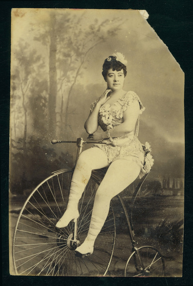 Woman in a leotard with white flowers, tights and heels, seated on a penny farthing bicycle.
