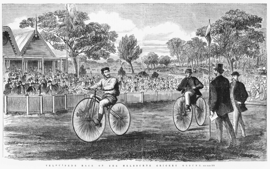 Black and white illustration of two men riding bicycles, with two men in coats and top hats beside. Behind a fence is a crowd of people with trees in the background.