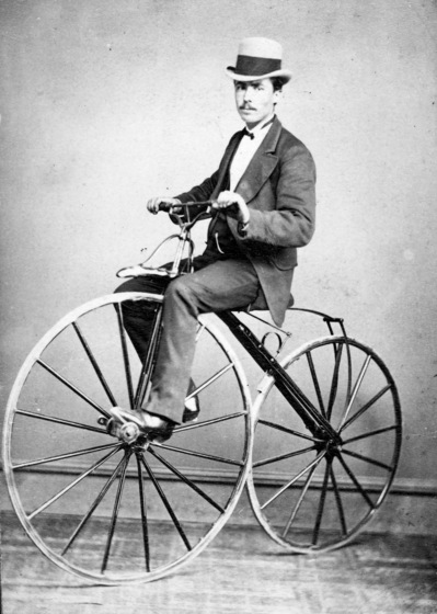 Man in a light coloured bowler hat and suit riding a bicycle