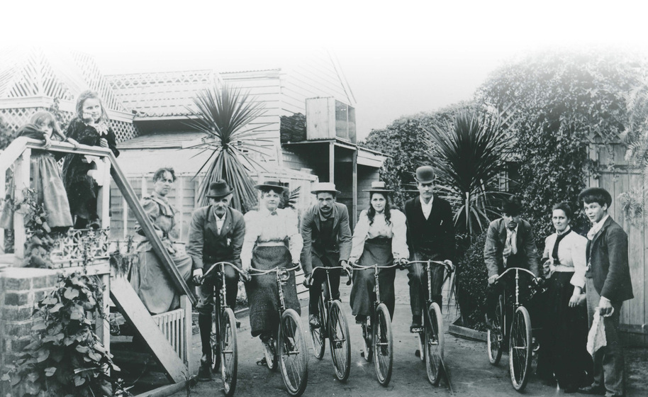 Four men and two women in 1890s clothing in a line on bicycles on a small town lane with buildings surrounding. People are standing either side, including two children on stairs.